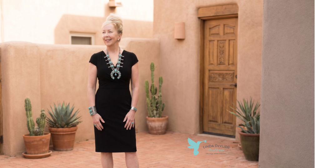 Professional woman in a black dress with turquoise jewelry posing in a sunlit adobe courtyard; real estate logo visible in the corner.